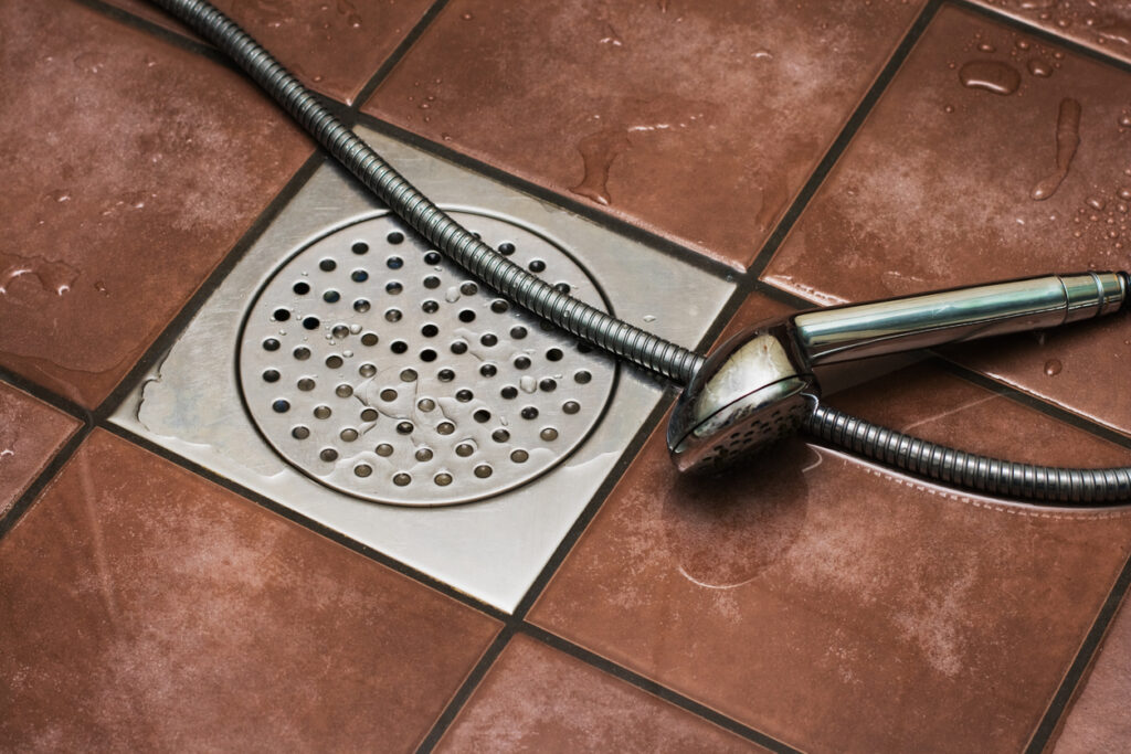 Detachable silver showerhead on floor of a shower with brown square tiles and square drain in the middle. Floor is wet.